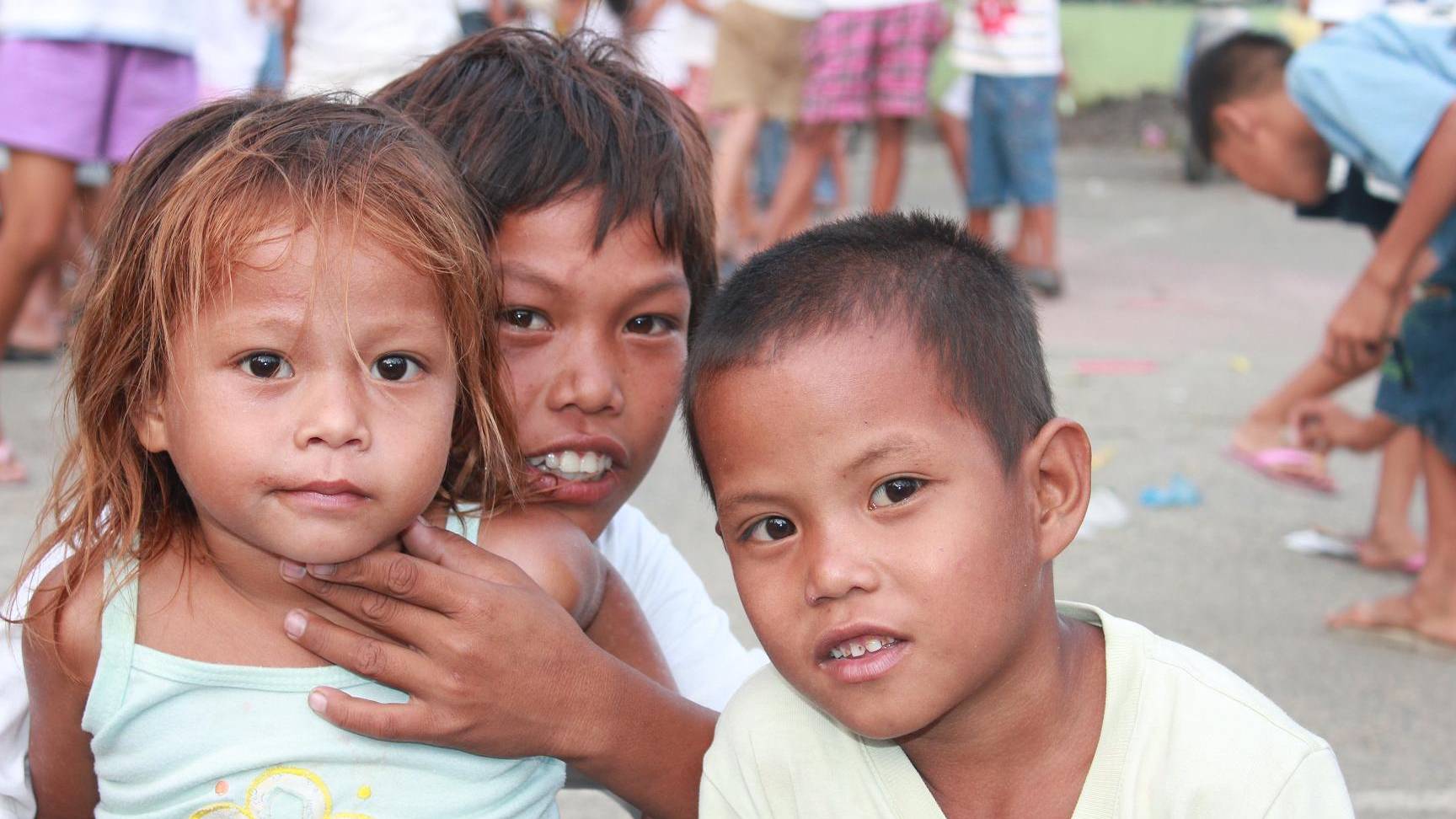 In the image, there is a group of children standing next to each other. The children are of different ages and genders, with a mix of boys and girls. One child is holding a toothbrush. Additionally, there is a man present, holding a baby. The children are wearing white shirts and appear to be in a crowded area with other people around. The children seem to be posing for a picture or simply standing together. The image captures a moment of innocence and togetherness among the group of children and the man with the baby.