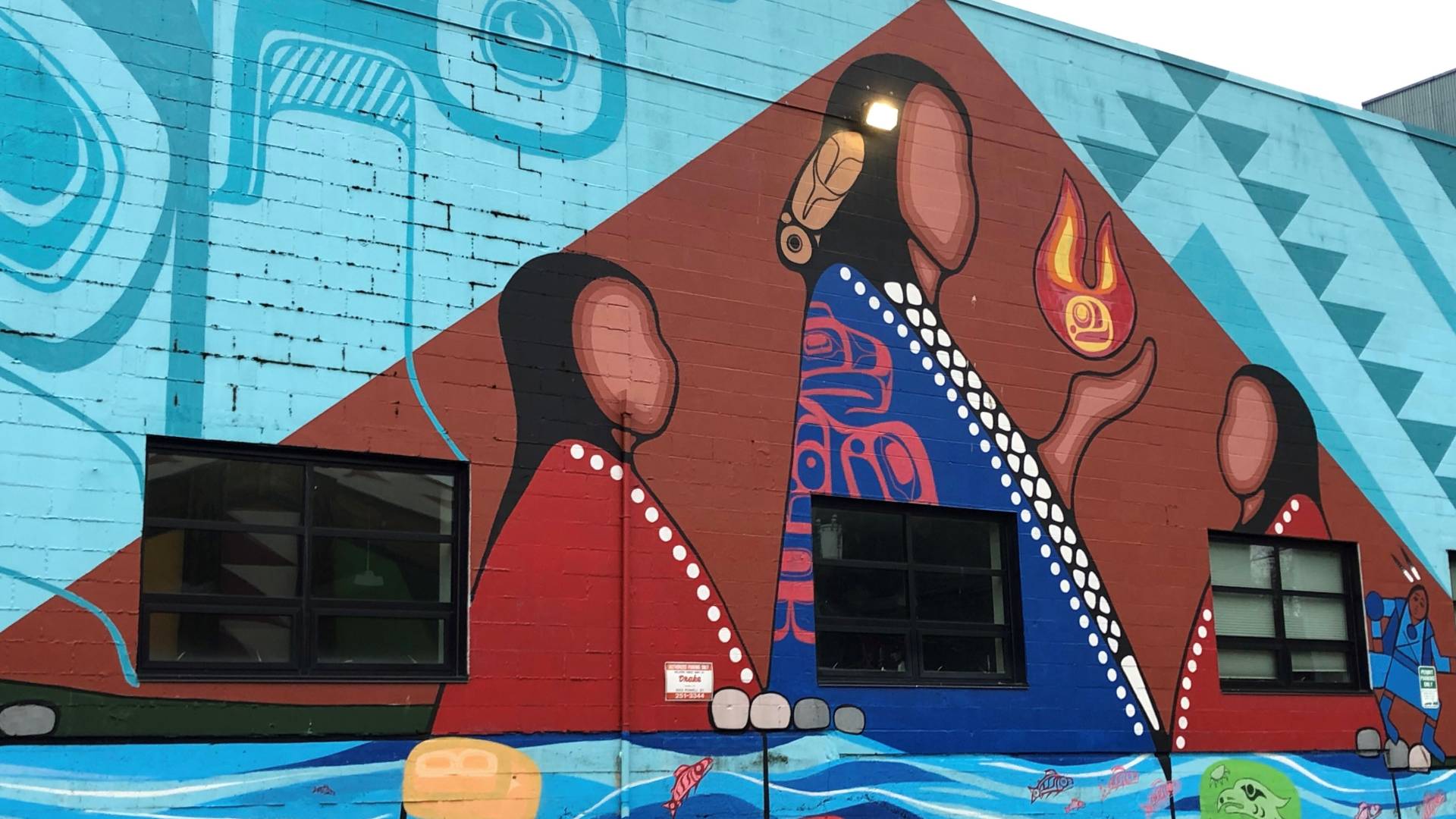 A beautiful mural adorns the side of a building, featuring a woman and a man. The woman is wearing a striking red dress, standing out against the vibrant blue background. The intricate doodle-style artwork showcases native people in a city setting. The colors of the mural include shades of blue, red, and white. The artwork is detailed and captivating, adding a burst of artistic flair to the urban landscape. This mural is a true work of art that captures the essence of cultural diversity and creativity.
