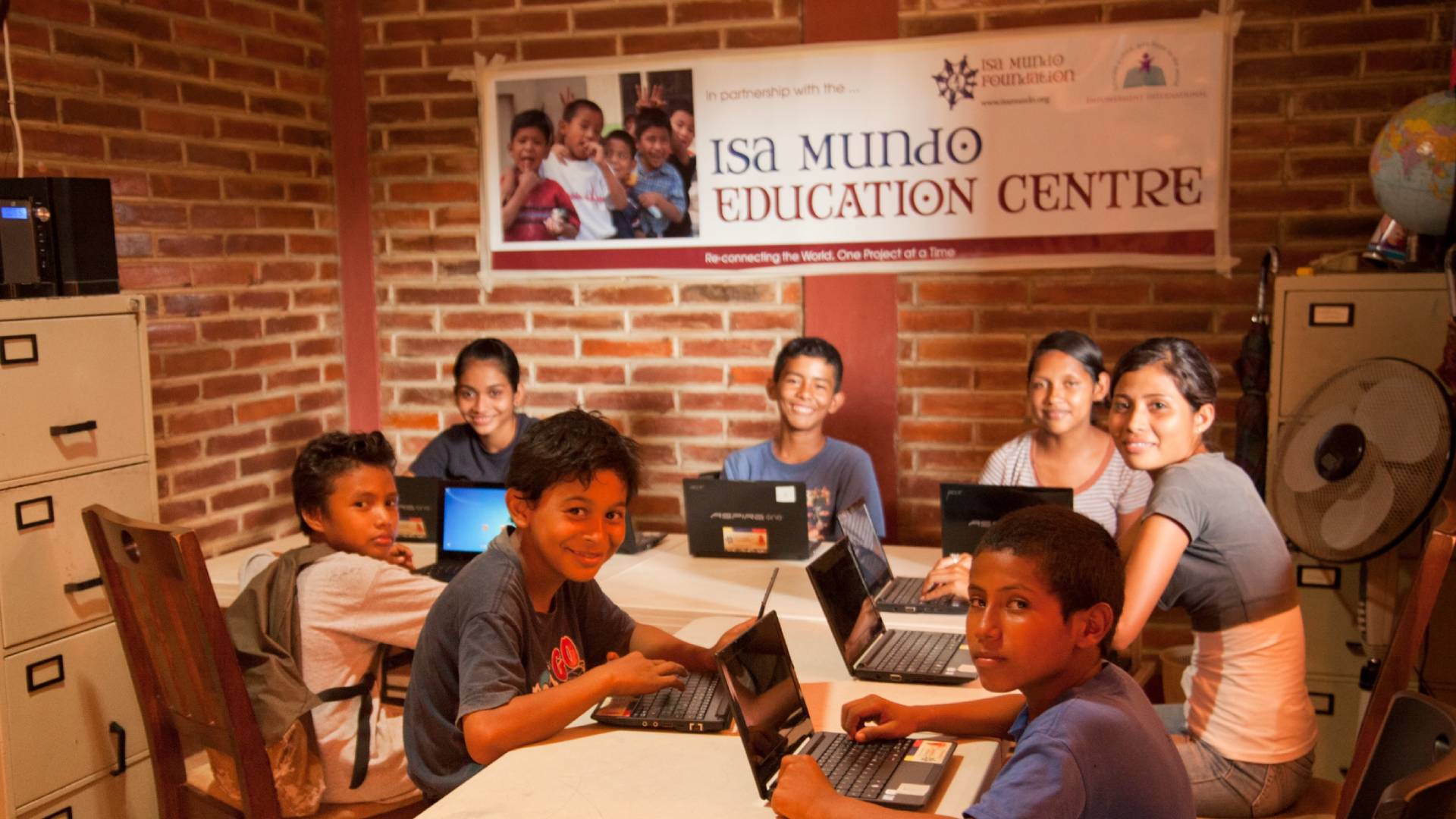 A group of children of various ages and genders are sitting at a table in what appears to be a classroom setting. They are all engaged with laptops, some smiling as they work. The scene is bustling with activity as the children focus on their screens. The room is bright with natural light, and there are chairs and a cabinet/shelf in the background. The children are surrounded by laptops, with some placed on the table and others on the floor. The image captures a moment of learning and collaboration as the children use technology for their educational activities.