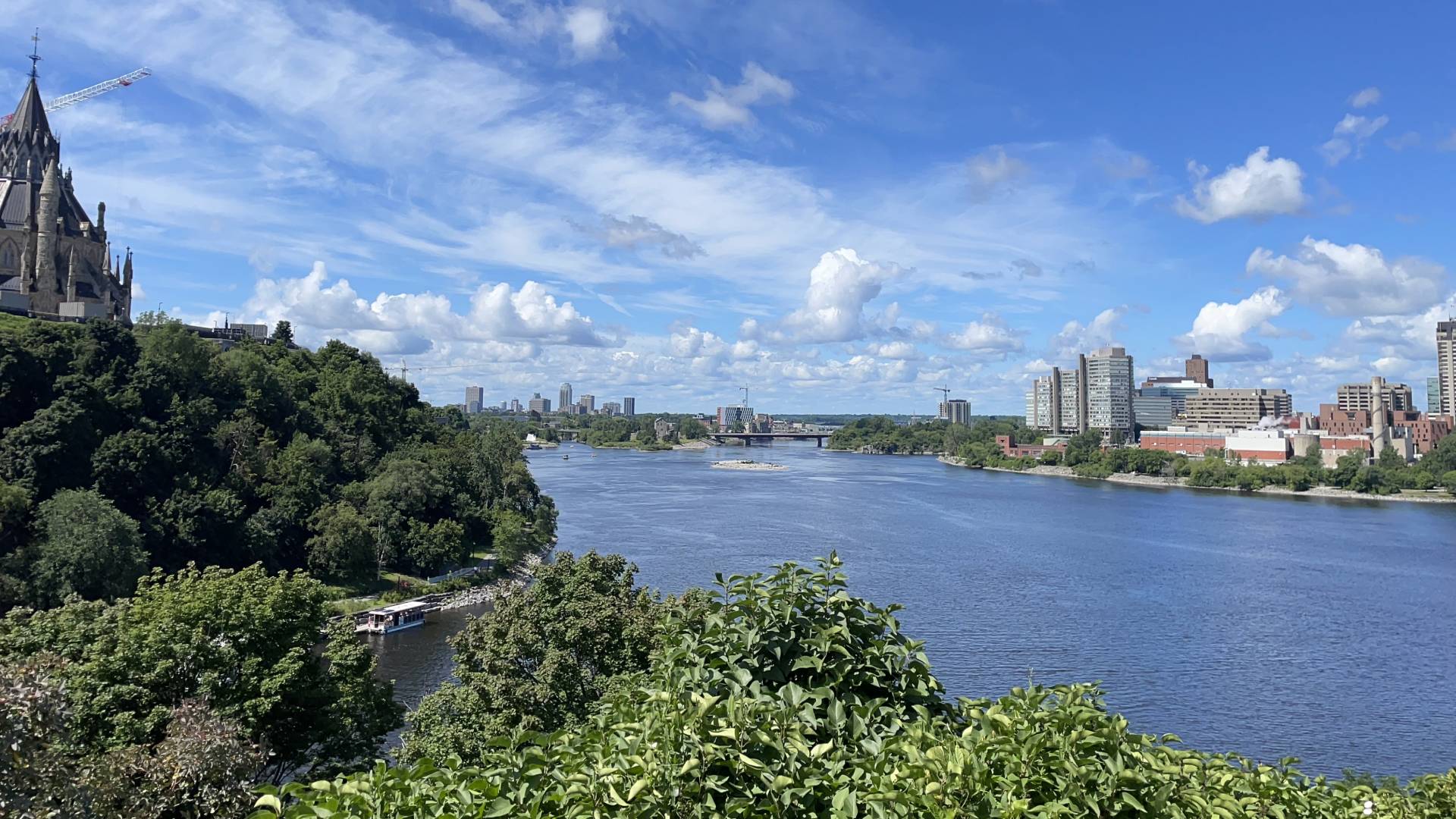 A picturesque scene of a serene river winding through a cityscape, with a majestic castle towering in the background. In the foreground, a tower crane stands tall against the sky, overseeing the bustling city below. Boats can be seen peacefully sailing along the river, adding to the charm of the setting. The city is dotted with buildings, factories, and greenery, creating a vibrant and lively atmosphere. The sky above is a calming shade of blue, completing the idyllic scene of urban beauty and natural tranquility blending seamlessly together.